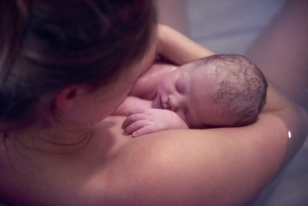 Over shoulder portrait of young woman holding new born baby daughter in birthing pool at home